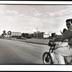 de la série "Isla" "Andris" portrait en demi hauteur et de profil d'un homme sur sa moto circulant sur une route bordée par un panneau de propagande communiste avec un portrait de Che Guevara